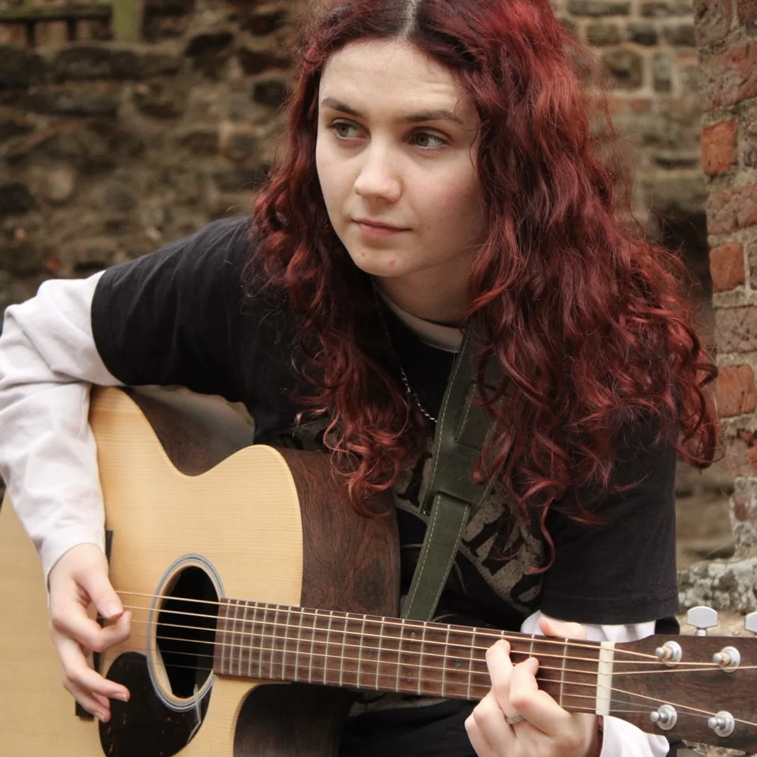 Grace Cope playing an acoustic guitar beside an old brick wall