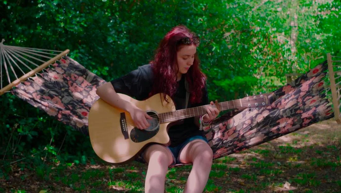 Grace Cope playing an acoustic guitar on a hammock among trees